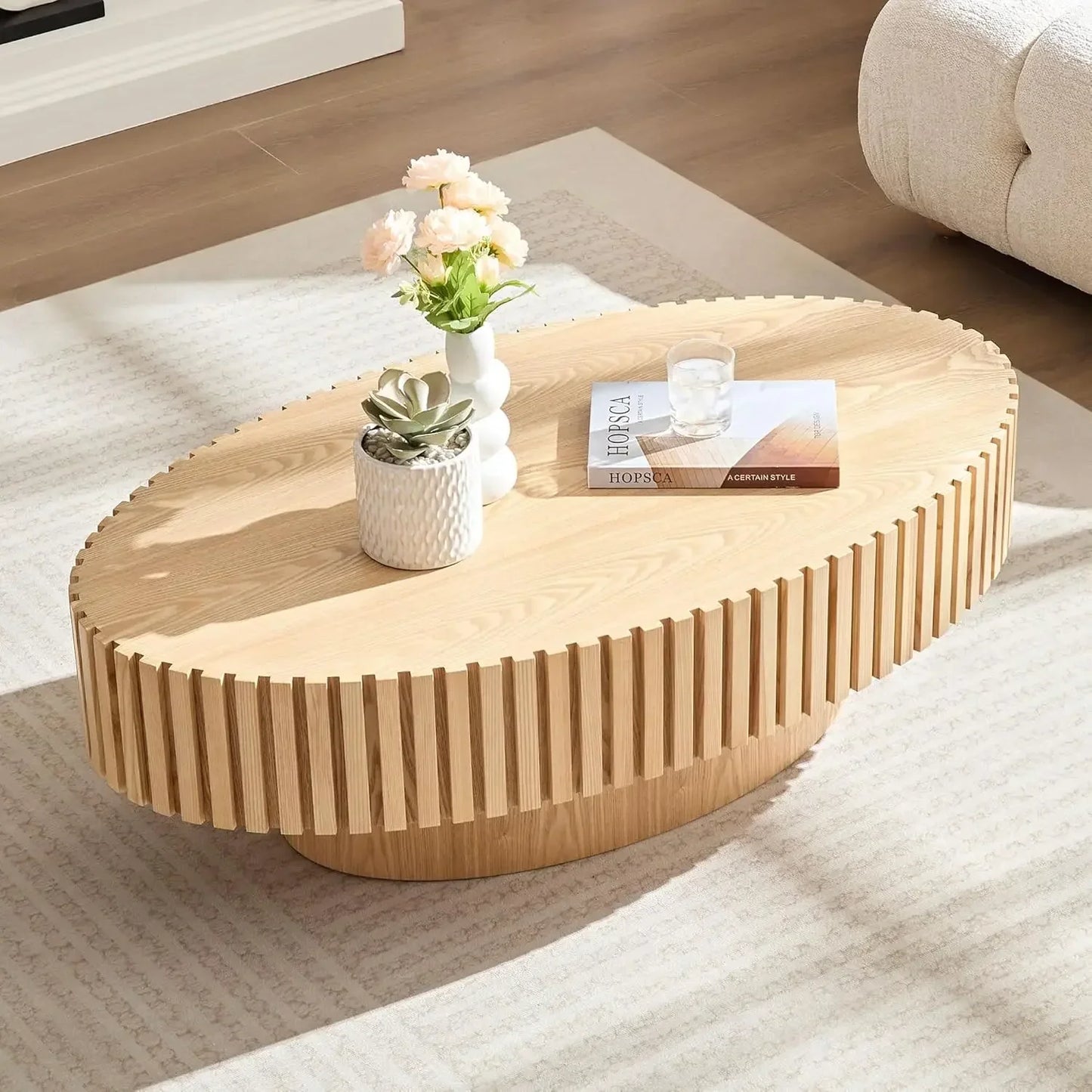 The 43" Morsale oval wooden coffee table with a ribbed edge sits elegantly on a textured rug. It displays a white vase with pink roses, a small succulent pot, a book, and a glass. A portion of a white sofa is visible in the background.