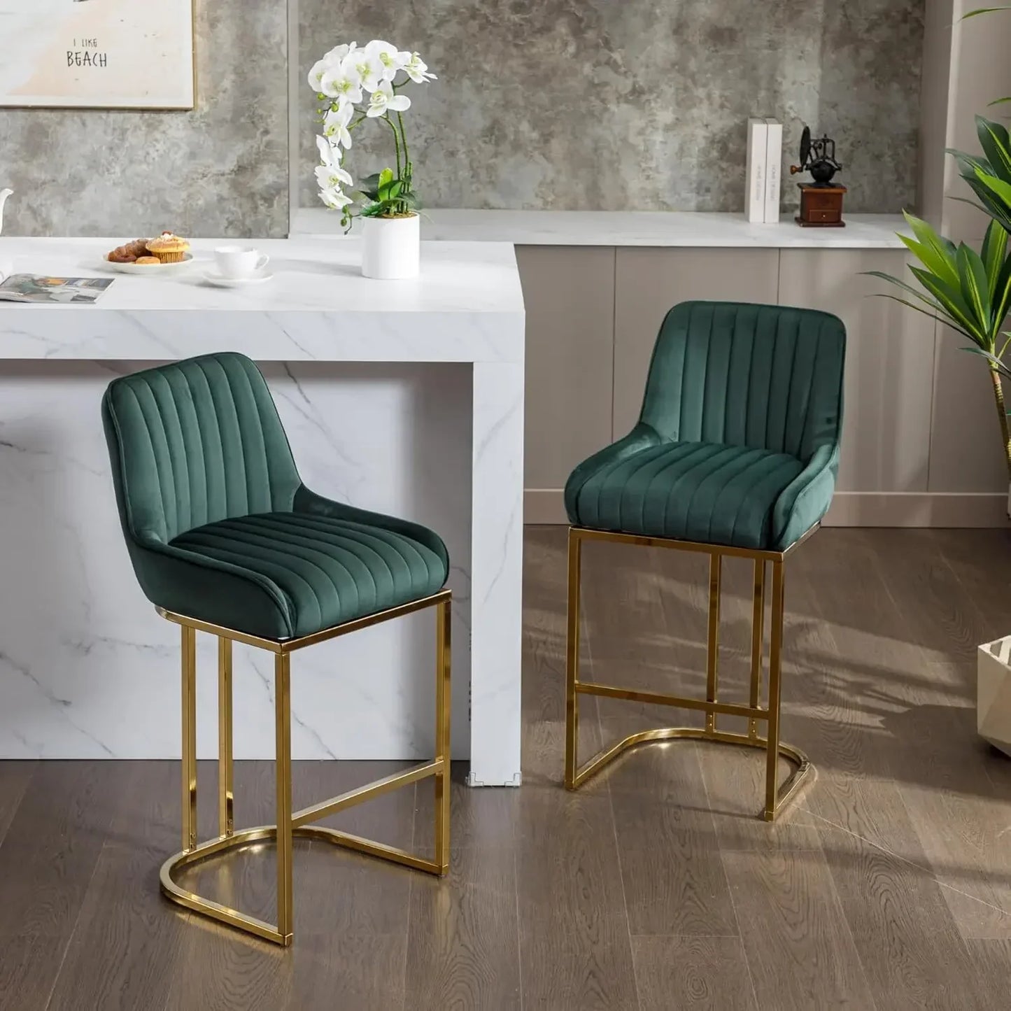 A set of two "Modern Counter Height Bar Stools" by Morsale, featuring green upholstery and a gold metal frame, complements a white marble kitchen island. A potted plant, an orchid on the counter, and a light gray textured wall complete the inviting atmosphere.