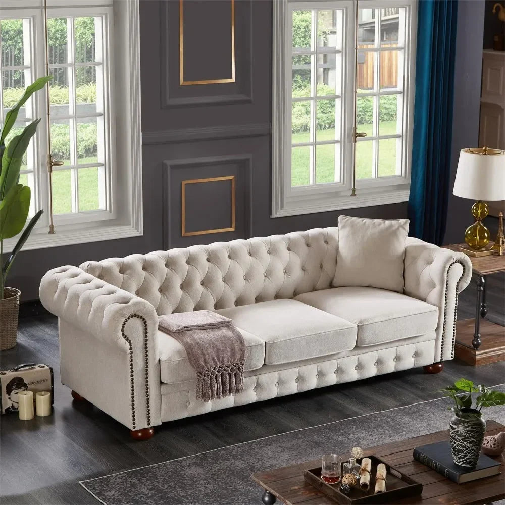 The living room showcases an elegant 3-Seat Chesterfield Sofa by Morsale, accented with a throw blanket and pillow. Dark wood flooring, potted plant, and large windows with a garden view complement the decor, which includes candles and a glass table for an inviting, stylish feel.