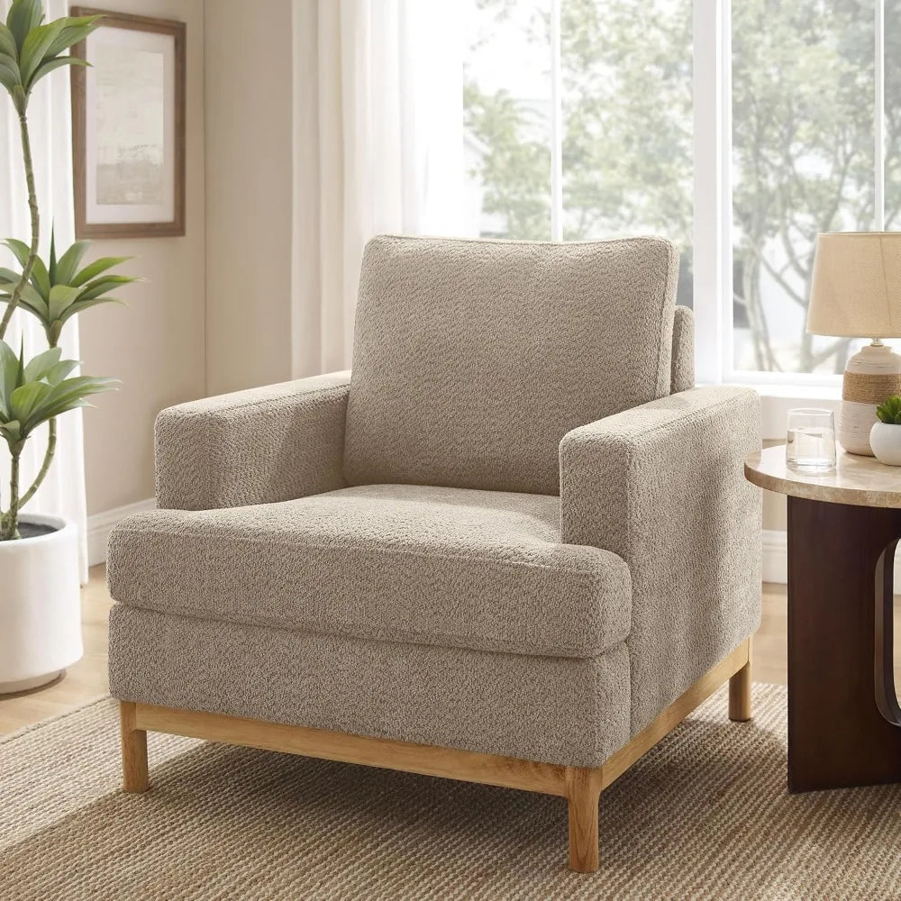 A Morsale Oversized Mid Century Modern armchair in beige, with wooden legs, is placed in a well-lit living room near a large window. The space includes a potted plant, wooden side table holding a lamp and glass of water, framed picture on the wall, and is adorned with a woven rug.
