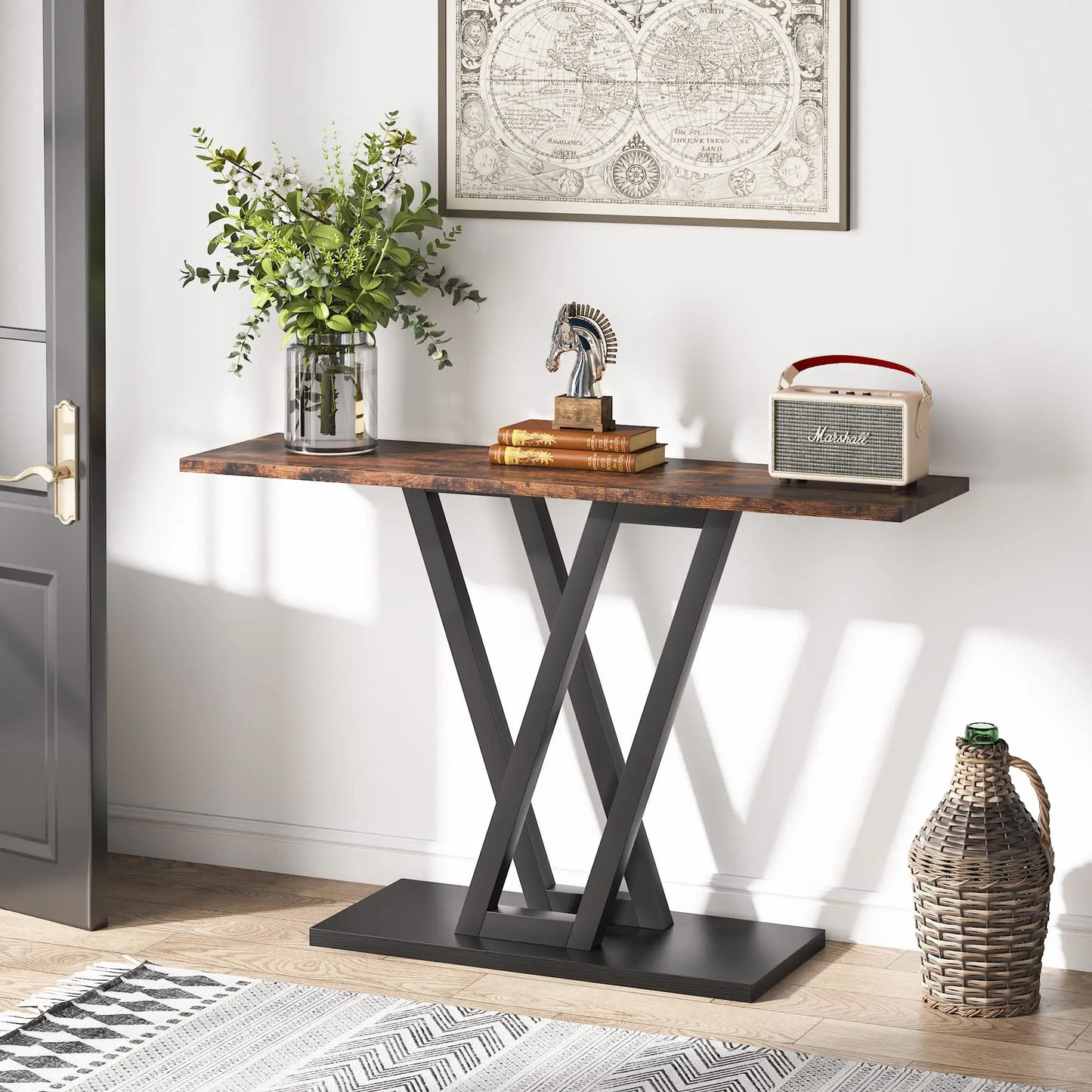 The Bradford Entryway Table by Morsale features a wooden top and sleek black crossed legs. It showcases a vase with a plant, a decorative sculpture, and a vintage radio beneath world map wall art. A wicker basket is placed on the floor nearby against the white wall, completing the living room setup.