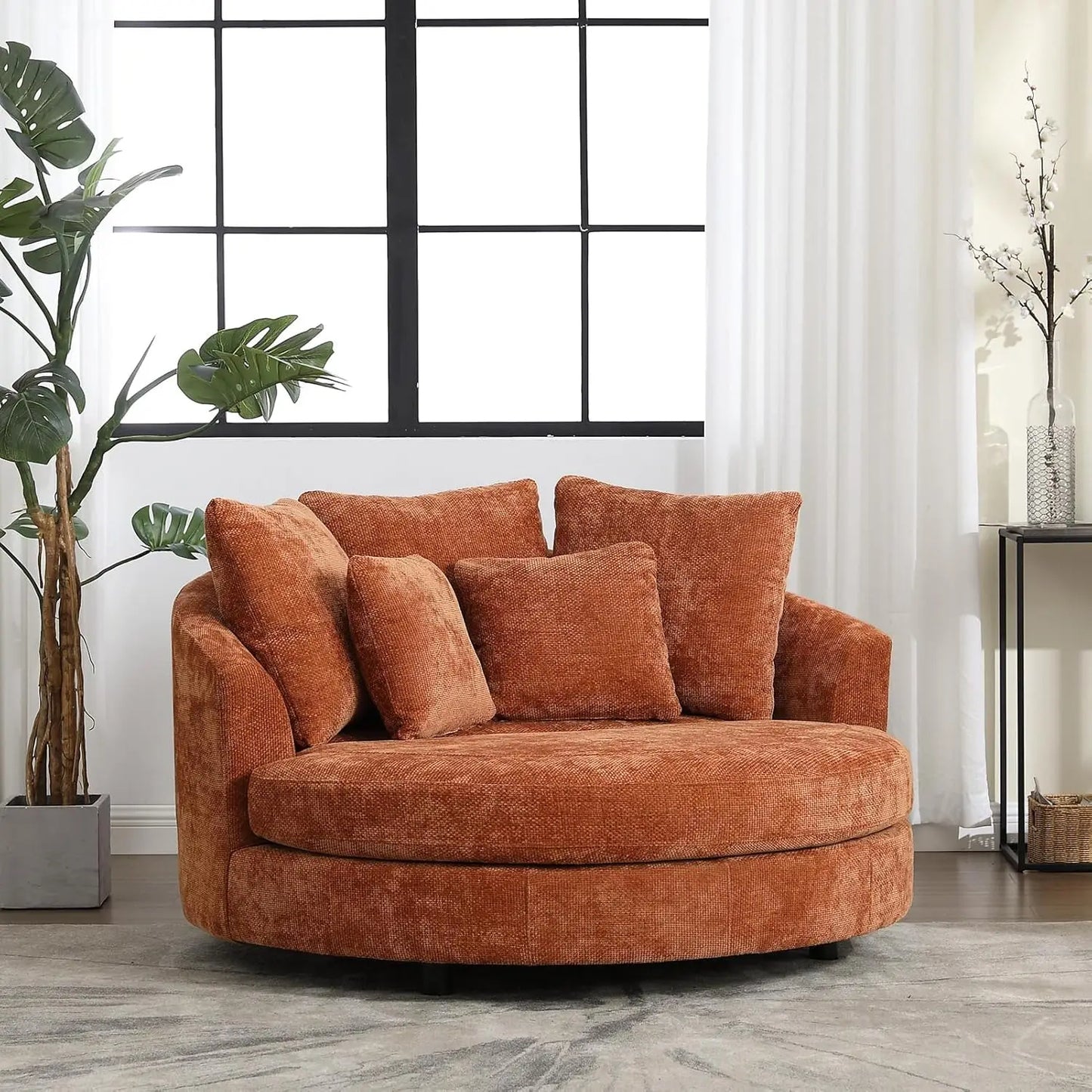In a well-lit room, Morsale's 59.8" Oversized Accent Barrel Sofa Chair in a cozy orange hue offers ample seating near a large window with sheer curtains, complemented by assorted cushions, a potted plant, a rug, and a small side table adorned with decor items.