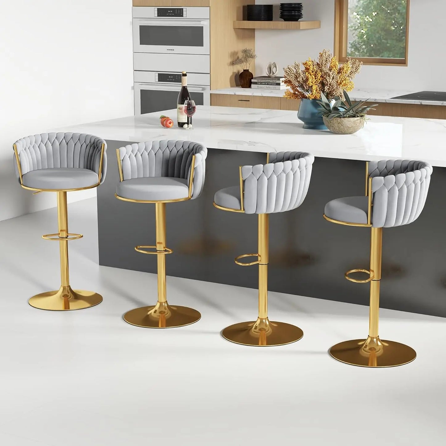 Four Morsale Height Adjustable Counter Stools with gold bases and gray quilted upholstery, featuring a 360° swivel, line up at a kitchen island. The kitchen has light wood cabinets, a white counter, built-in ovens, and a window adorned with a wine bottle and flower arrangement.