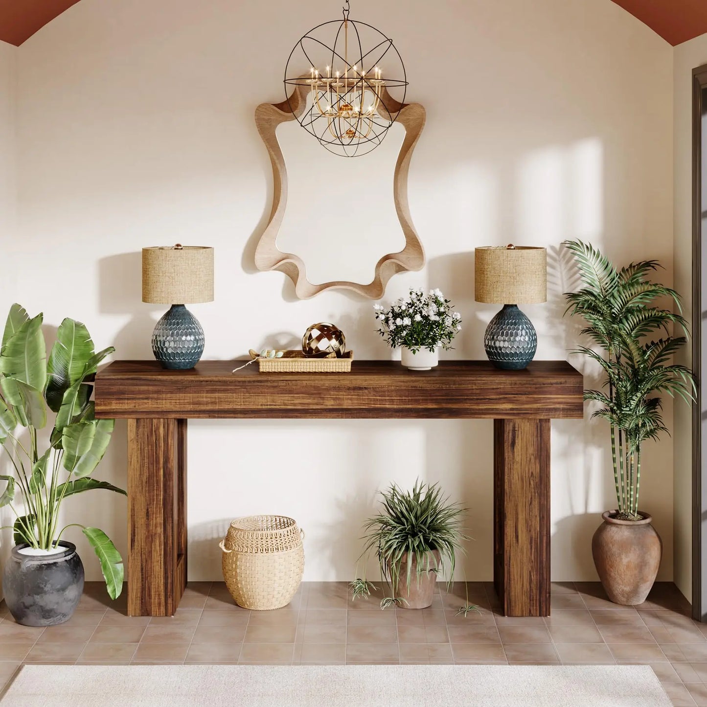 A Farmhouse 63-Inch Entryway Console Table by Morsale rests against a light wall, adorned with two blue lamps and decor. A wavy-edged mirror above adds retro charm, while antique potted plants provide greenery to the tiled floor. Overhead, a chandelier completes the scene.