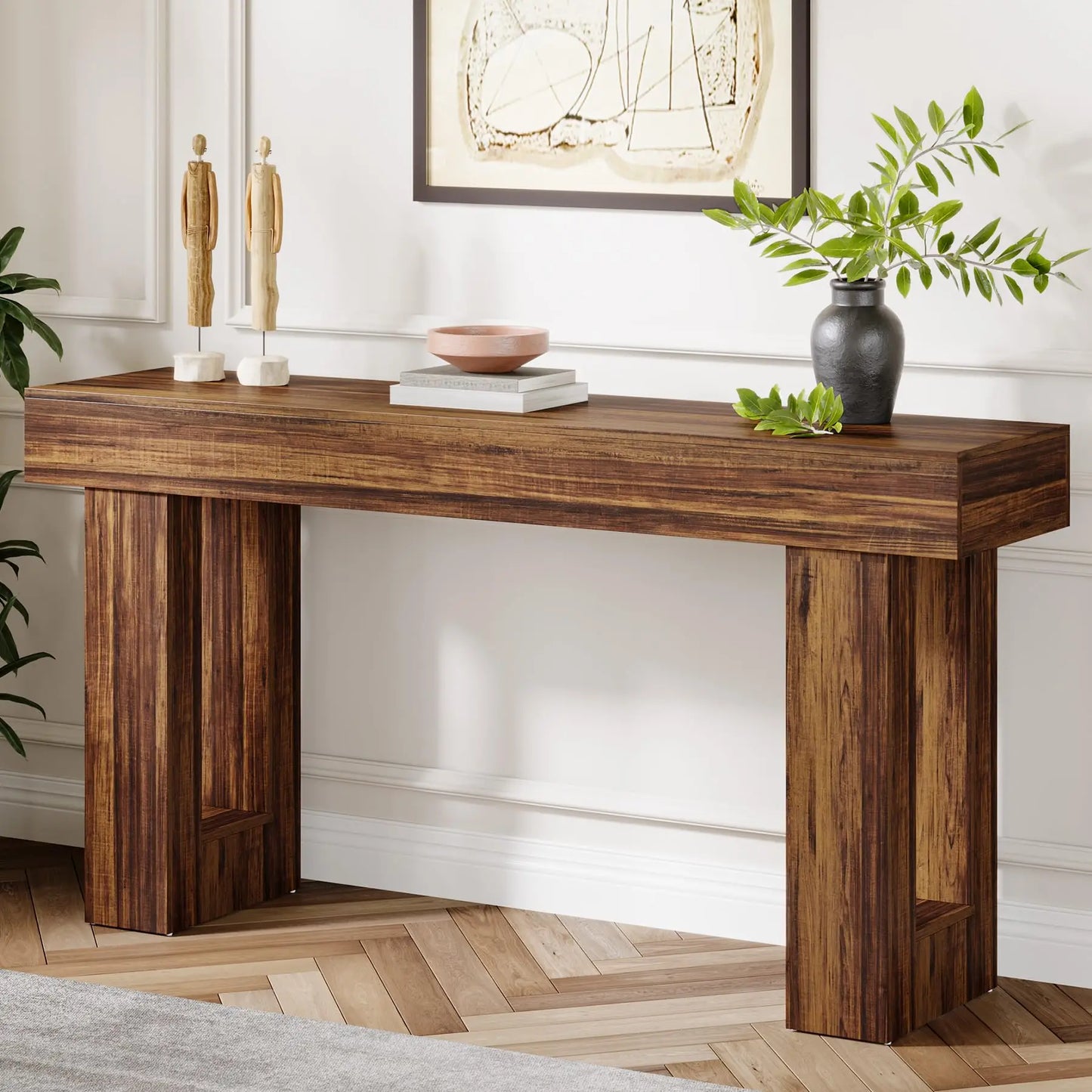 The Morsale Farmhouse 63-Inch Entryway Console Table, with its rich brown finish, stands elegantly against a white wall. It holds two small sculptures, a peach-colored bowl, and a black vase with green branches. Above it hangs framed abstract art on the chevron-patterned wooden floor.