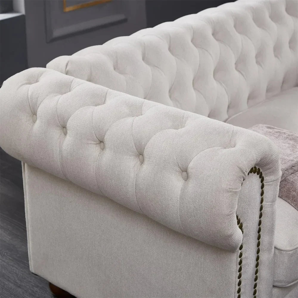 A close-up of the tufted beige armrest on Morsale's 3-Seat Chesterfield Sofa reveals button detailing and nailhead trim. It rests on a dark wooden floor with a glimpse of a gray wall in the background.