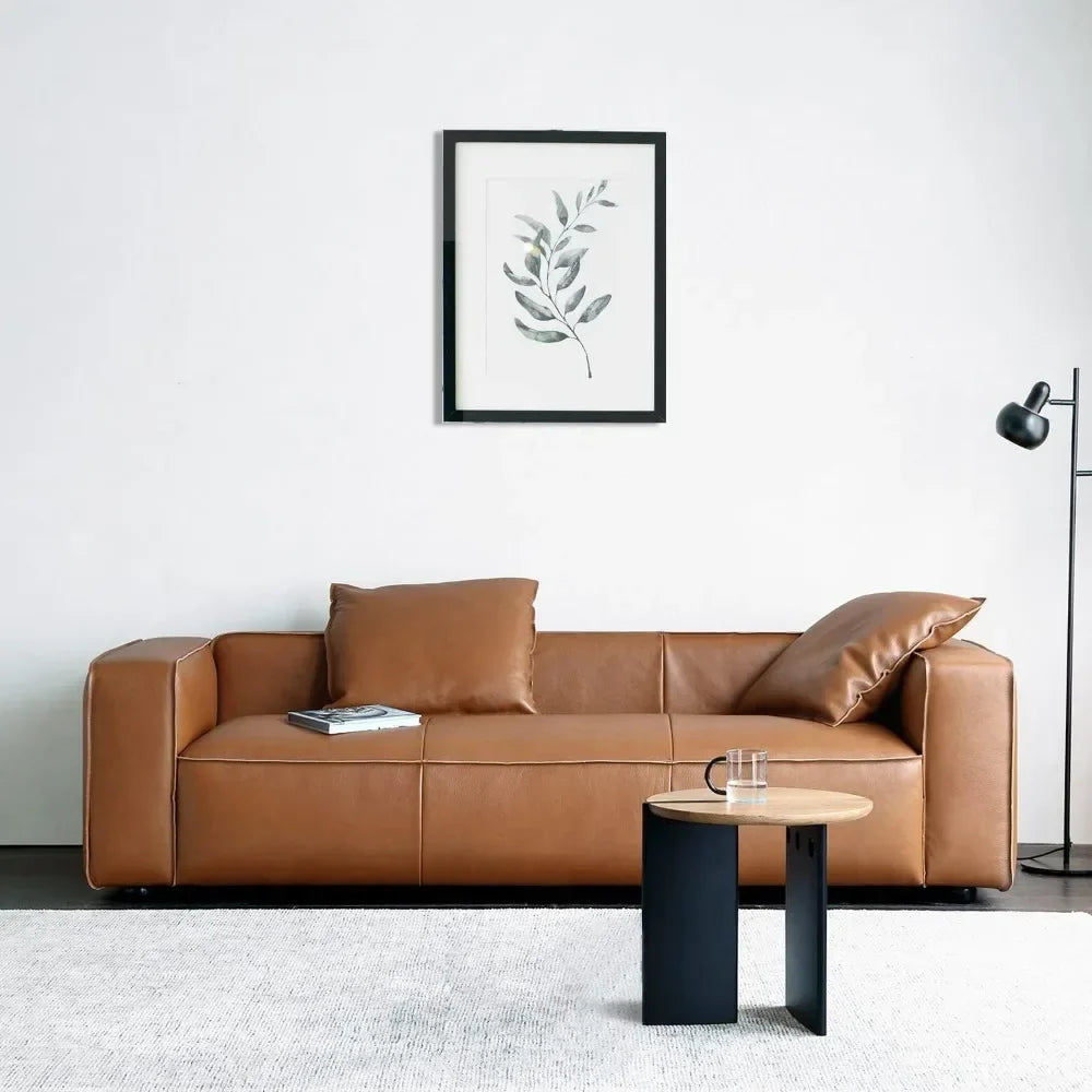A minimalist living room showcases Morsale's Stefano Genuine Leather Lounge Sofa as the centerpiece, paired with a small round side table holding a glass and mug, and a sleek standing lamp. A framed botanical print decorates the white wall, while a light-colored rug completes the modern look.