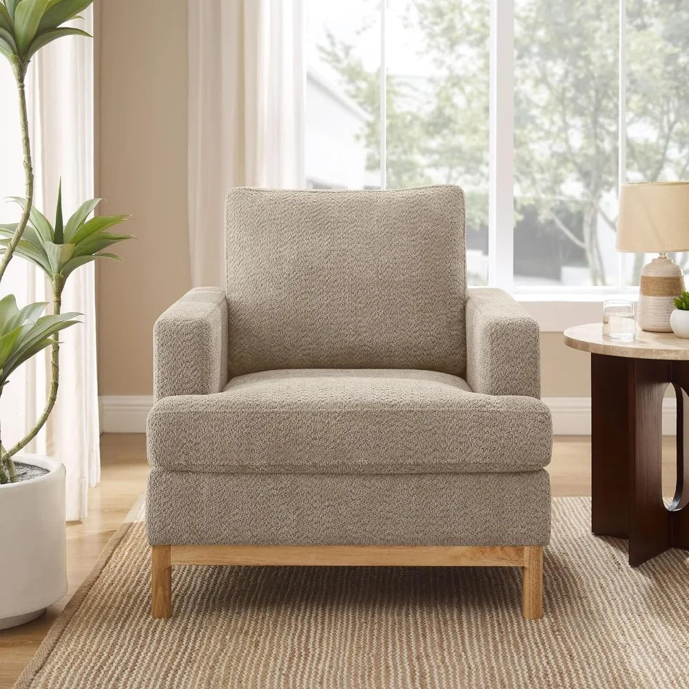 A Morsale Oversized Mid Century Modern beige armchair with wooden legs sits on a striped area rug, accompanied by a wooden side table holding a lamp and decor items. A large window showcases greenery outside, while a tall plant nearby adds to the room's cozy ambiance.