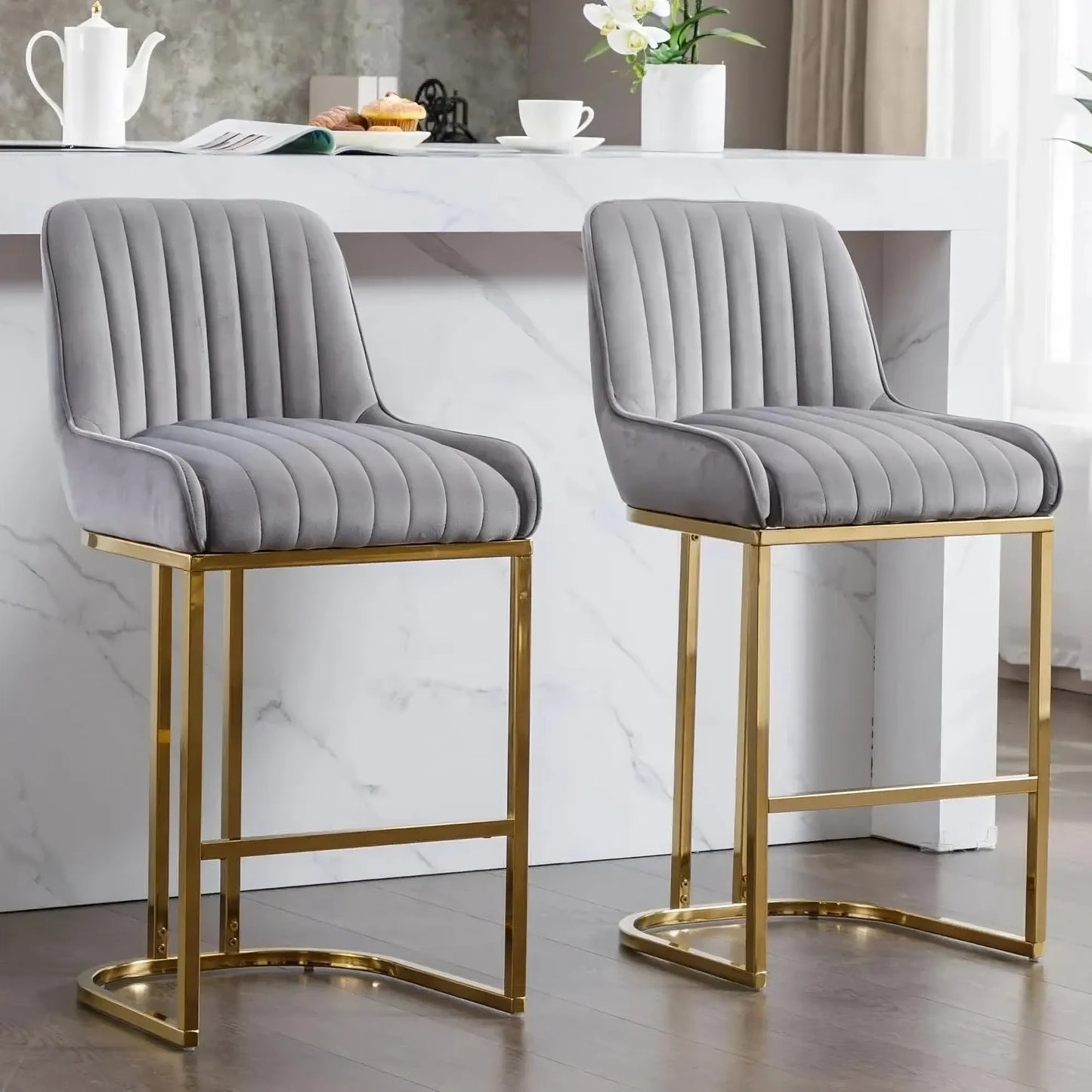 A set of two Morsale Modern Counter Height Bar Stools, featuring gray vertically-stitched velvet seats and gold metal frames, pairs with a teapot, cup, and decor on the marble kitchen island as light streams in from the right window. Made in the US.