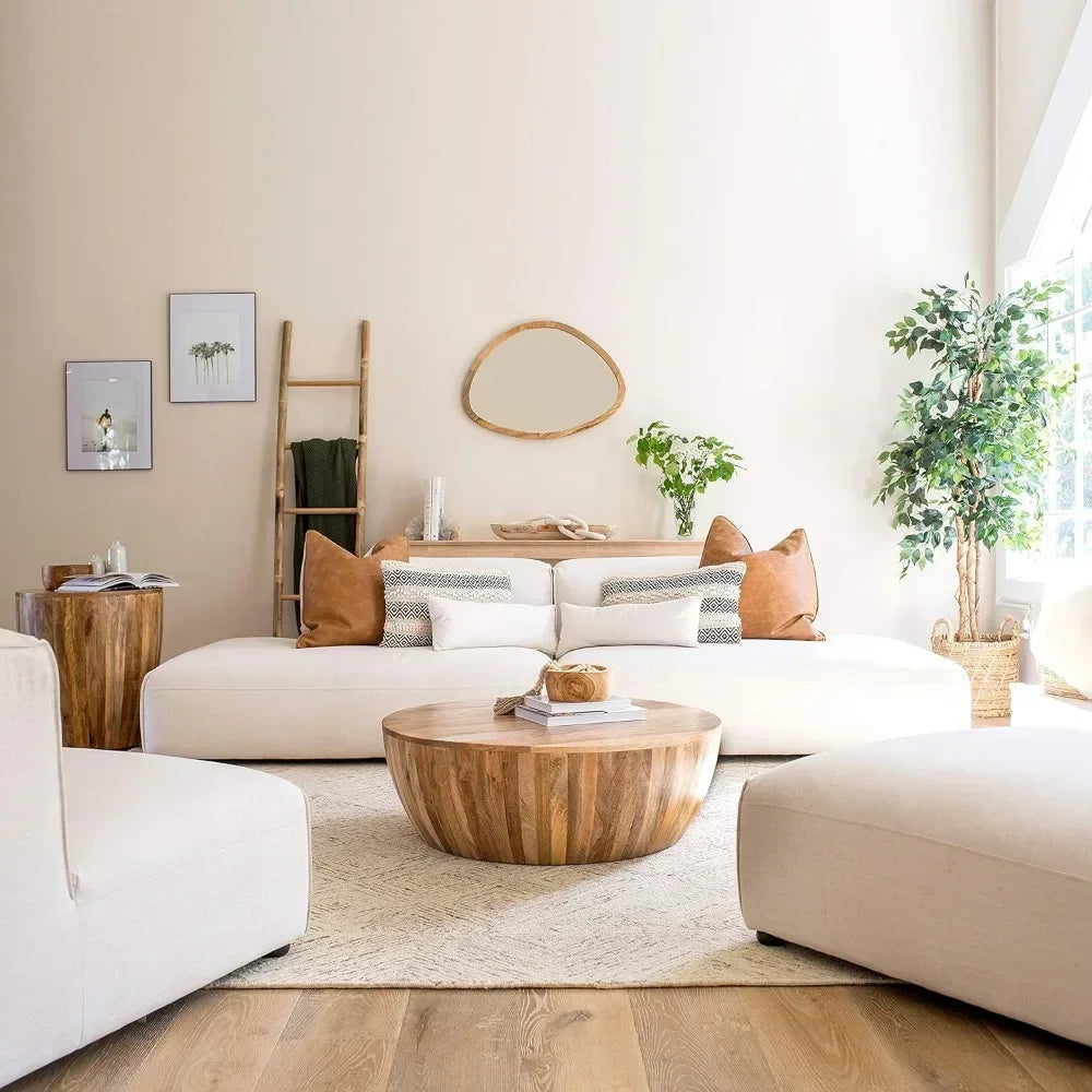 A cozy living room showcases a large white couch with brown pillows in a modern style. The space is anchored by a Morsale Mango Wood Goa Coffee Table and soft rug. A ladder shelf with decor, wall art pieces, and a large plant by the window complete this inviting scene.