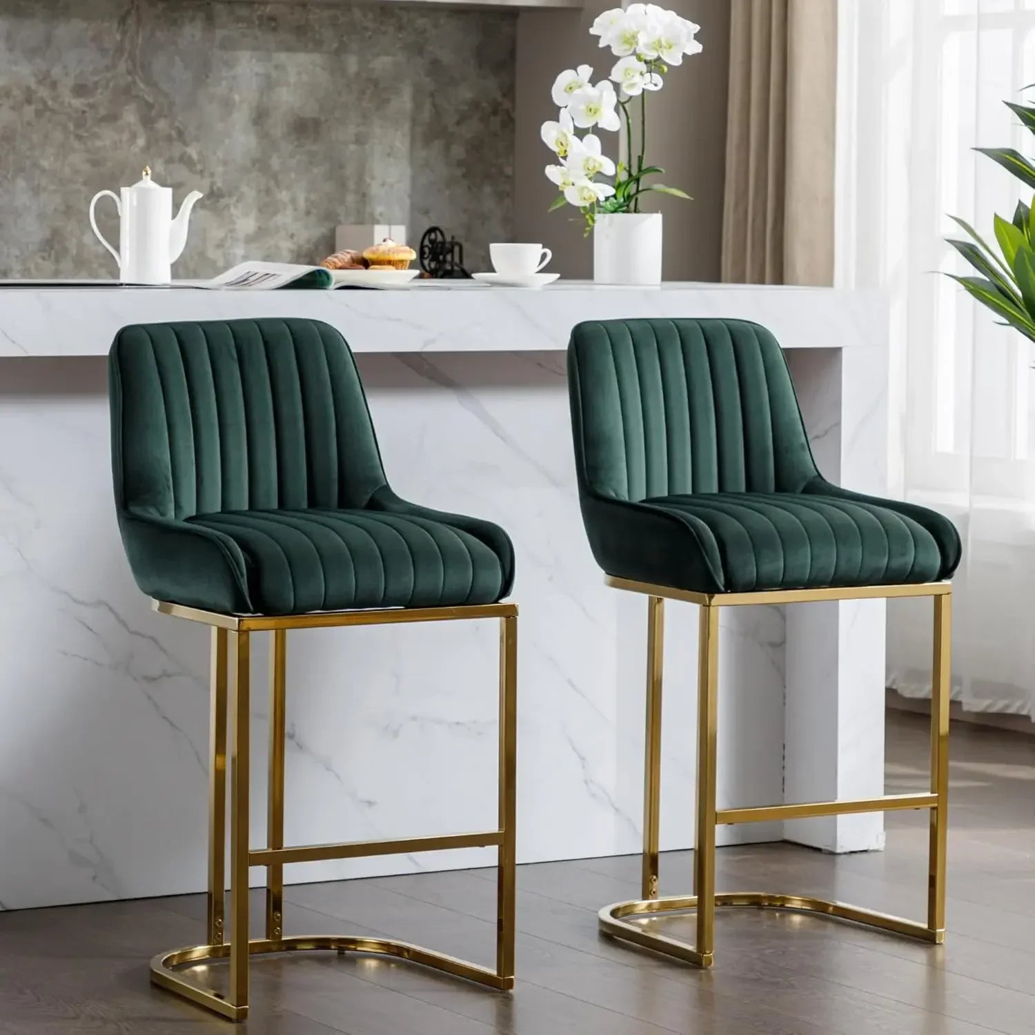 Two Morsale Modern Counter Height Bar Stools in green velvet with gold metal frames are in front of a marble kitchen island, where a white teapot and two cups sit alongside a potted orchid. The background shows a gray textured wall and a window with beige curtains.