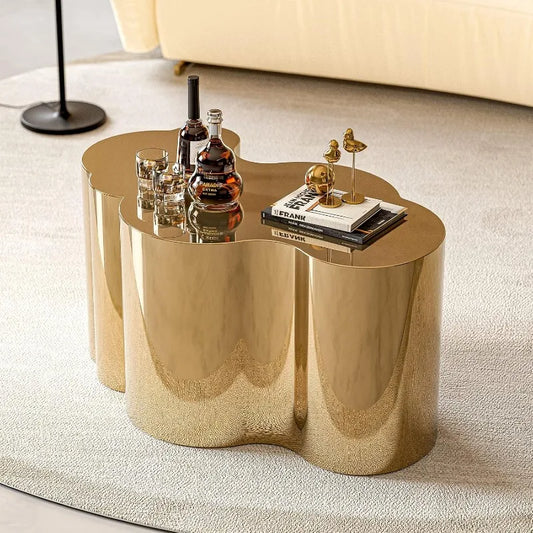 The Morsale Stainless Steel Gold Coffee Table sits on a light rug, adorned with a bottle, two glasses, books, and a small decorative piece. A beige sofa and sleek black floor lamp can be seen partially in the background.