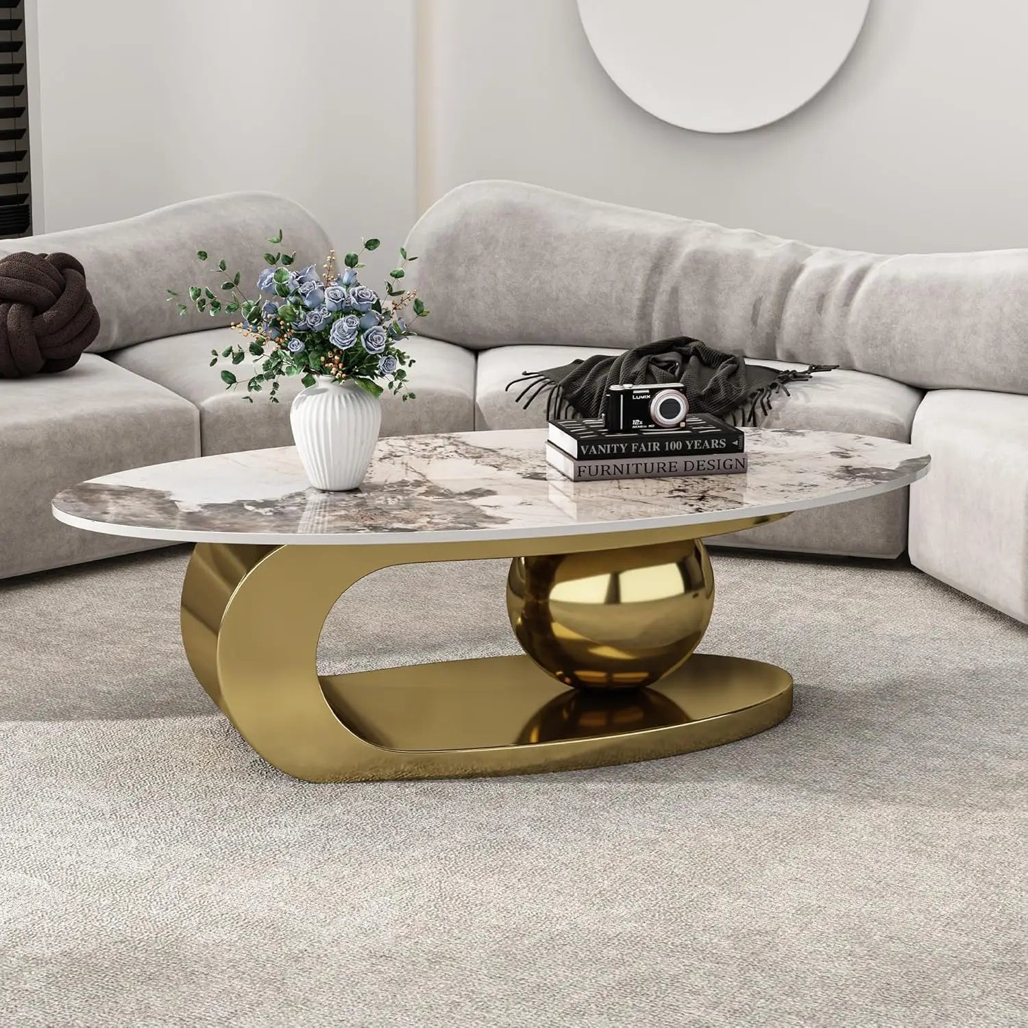 A modern living room displays a luxurious Morsale Modern Coffee Table with a premium ceramic top and unique gold oval steel base, highlighted by a large gold sphere. A white vase with blue flowers and a camera-themed magazine sit stylishly on the table, framed by a light gray sofa.