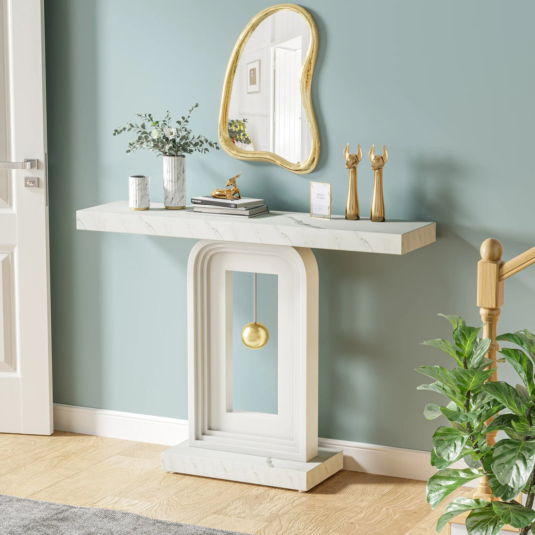 A chic, modern entryway features the Westford Contemporary Entryway Table by Morsale, highlighted by a gold pendulum accent. Decorative plants and gold figurines sit on top, while a wavy-edged mirror hangs above the minimalist teal wall. A potted plant and sleek wooden accents complete the look.