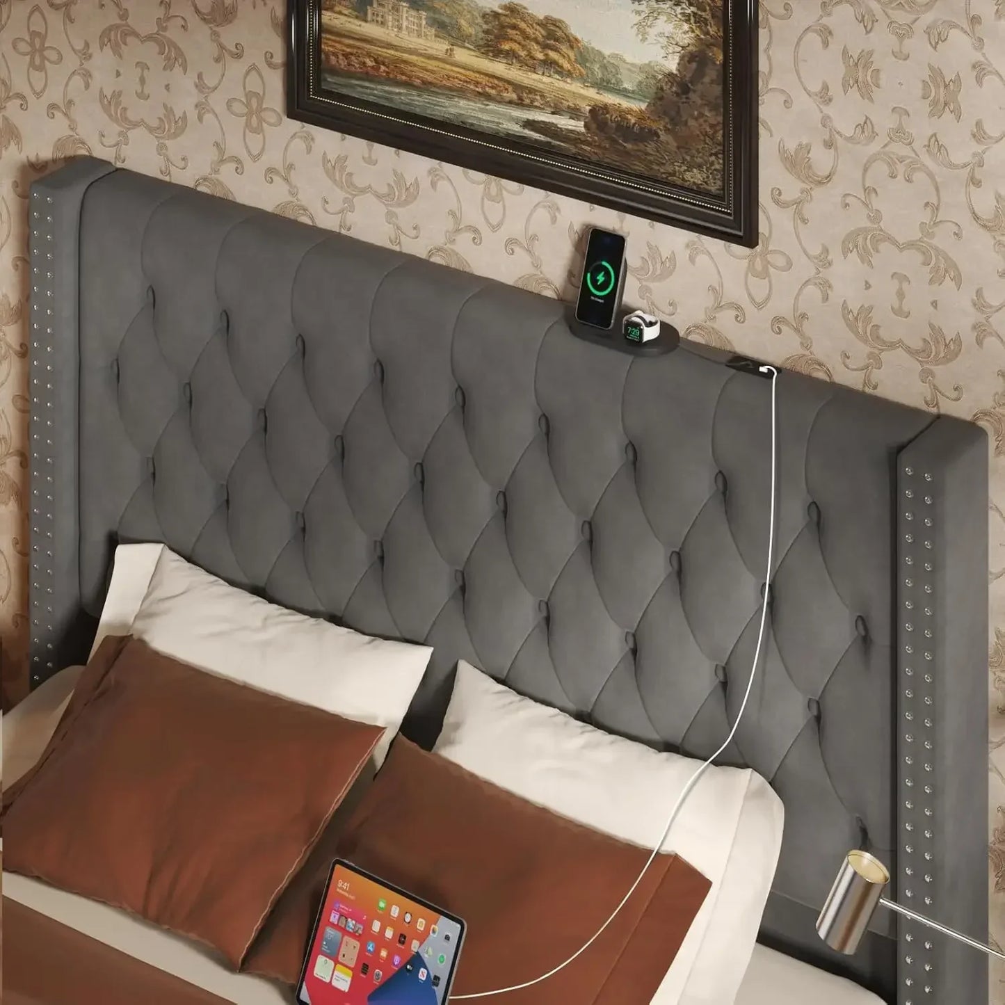 A bedroom scene showcases the Morsale Gio Velvet Queen Bed with a gray upholstered headboard, diamond tufting, and silver studs. Brown and white pillows rest on the bed. A tablet uses built-in charging ports on the headboard, with a framed landscape painting above.