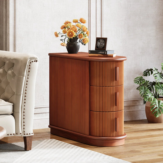 The Morsale Galway Solid Wood End Table, with its vertical grooves and three drawers, exudes rustic charm. Featuring a vase of orange flowers, a framed photo, and two books, it complements the room next to a beige tufted armchair on a wooden floor beside a large potted plant.