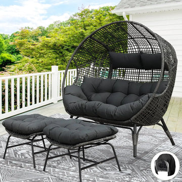 The Morsale Winsford Patio Lounge Chair in black PE wicker, featuring round cushions and crafted with weather-resistant fabric, sits on a deck amidst greenery. Two matching footrests rest on a patterned outdoor rug. A white railing and part of the house are visible.