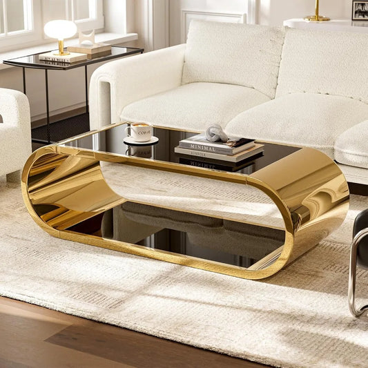 In a stylish living room, the Morsale Stainless Steel & Glass Coffee Table features a modern gold and black oval design with a reflective surface. Adorned with books and a coffee cup, it's complemented by a white textured sofa and glass-top side table for an elegant setting.