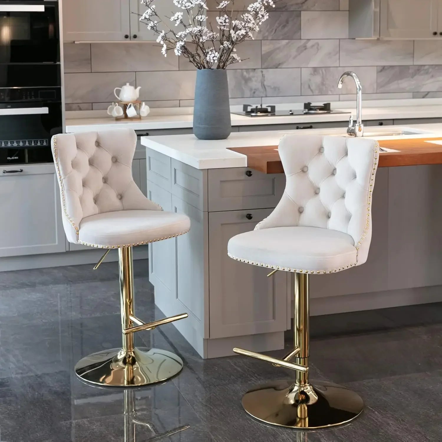 In the modern kitchen, a set of two elegant Morsale Counter Height Swivel Bar Stools with ergonomic tufted white upholstery and gold bases enhances the space. They offer adjustable height swivel function and complement the gray cabinets, marble backsplash, and central island with white flower vase.