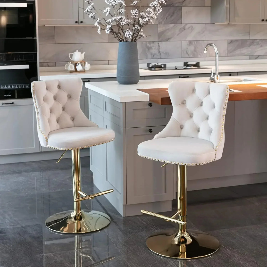 In the modern kitchen, a set of two elegant Morsale Counter Height Swivel Bar Stools with ergonomic tufted white upholstery and gold bases enhances the space. They offer adjustable height swivel function and complement the gray cabinets, marble backsplash, and central island with white flower vase.