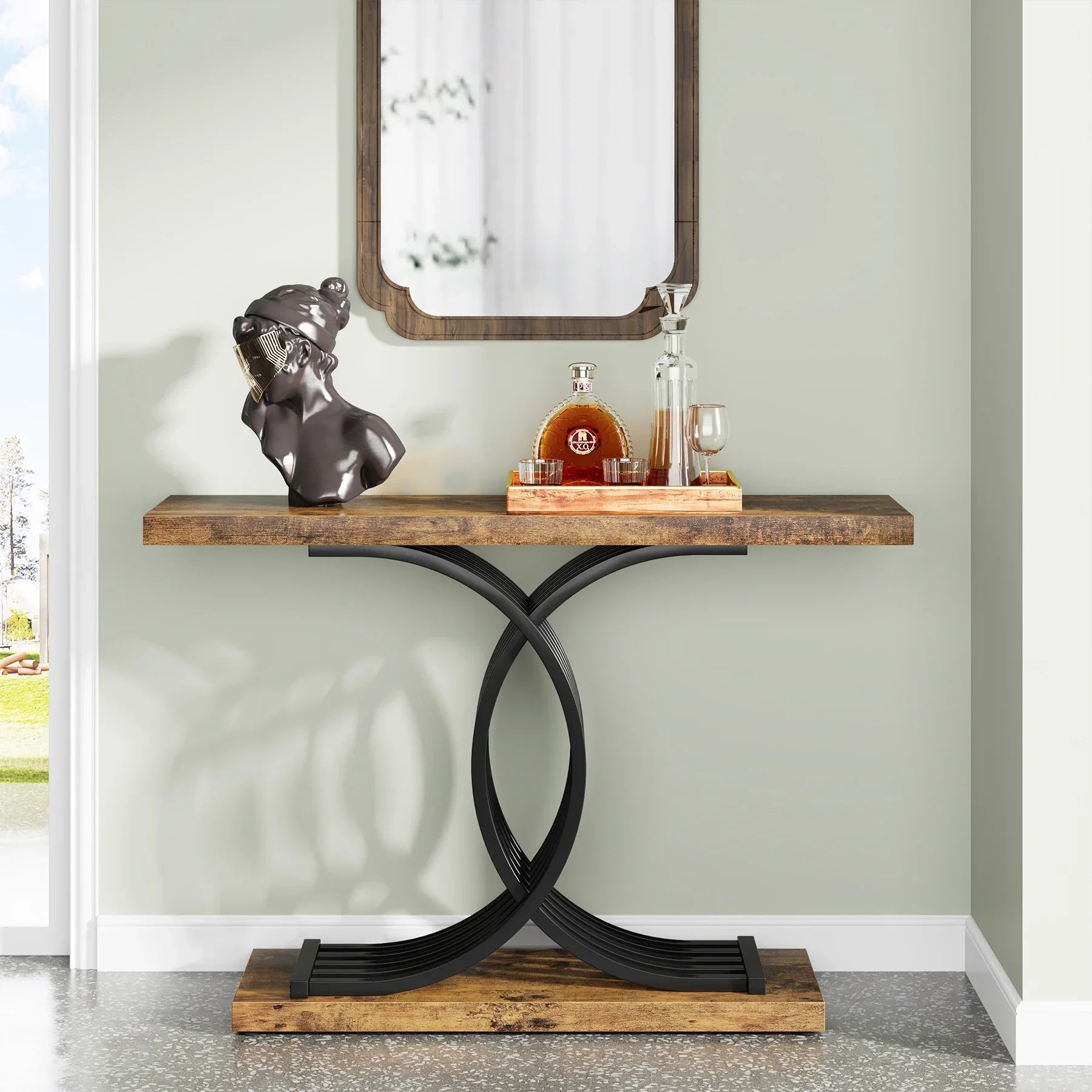 The Wexford Geometric Frame Console Table by Morsale features a rustic wooden top and modern black, intertwined metal legs. On its rectangular surface sits a decorative sculpture, a glass decanter with amber liquid, and two glasses on a tray, complemented by a mirror above.