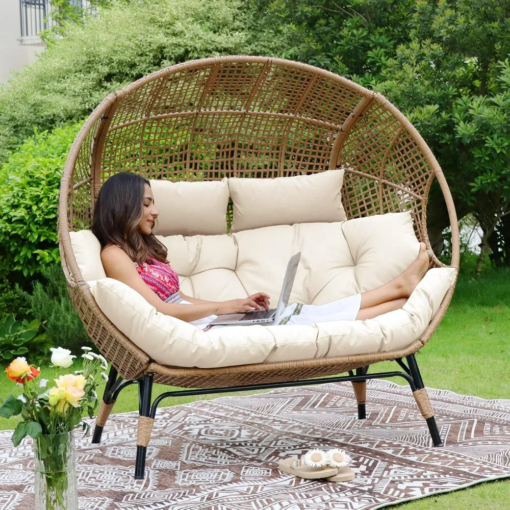 The Morsale Winsford Patio Lounge Chair in black PE wicker, featuring round cushions and crafted with weather-resistant fabric, sits on a deck amidst greenery. Two matching footrests rest on a patterned outdoor rug. A white railing and part of the house are visible.