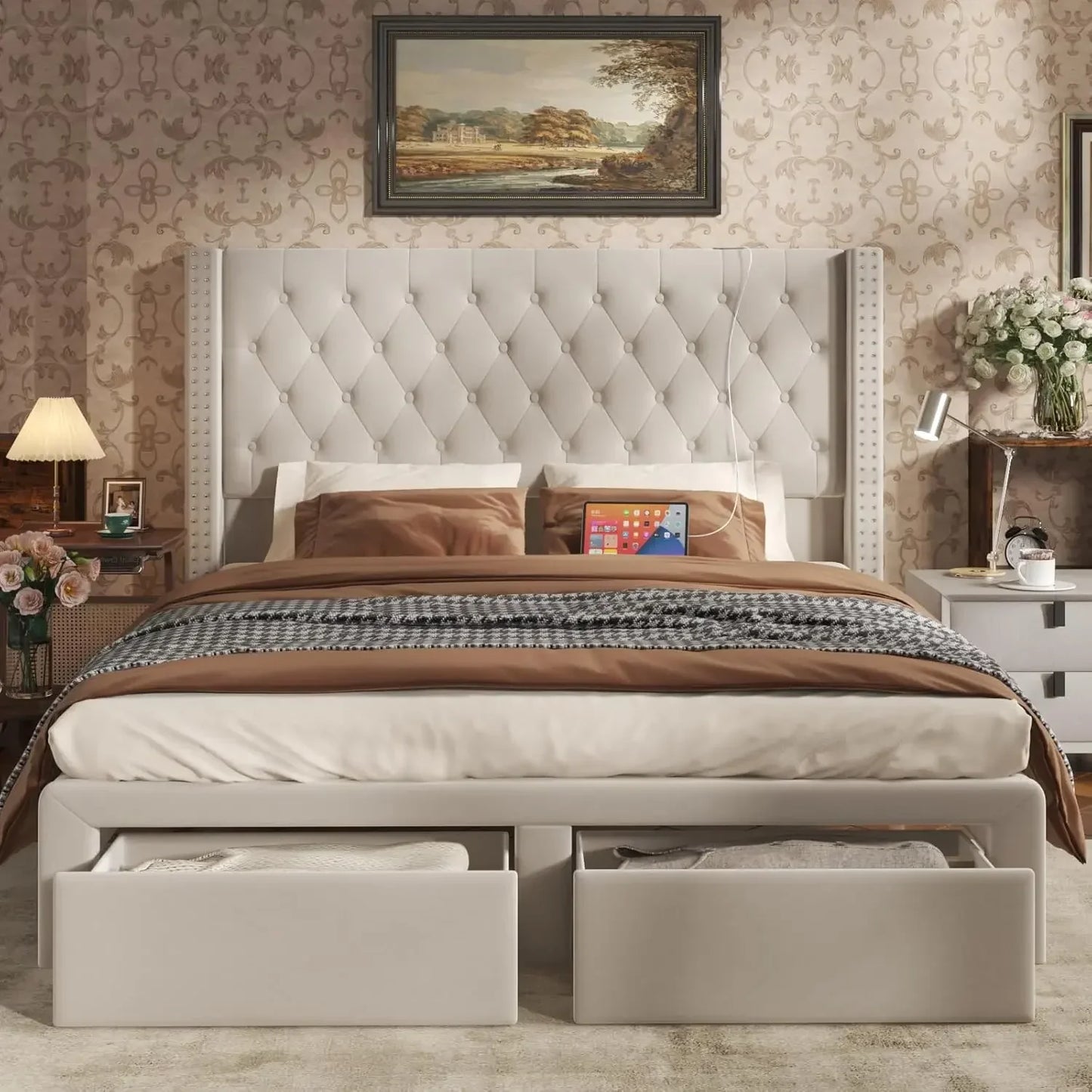 The Gio Velvet Queen Bed with Storage by Morsale creates a luxurious bedroom with its modern tufted headboard. Brown and beige cotton bedding complements elegant patterned wallpaper, while two storage drawers, a painting above, and bedside tables with lamps and flowers enhance the cozy ambience.