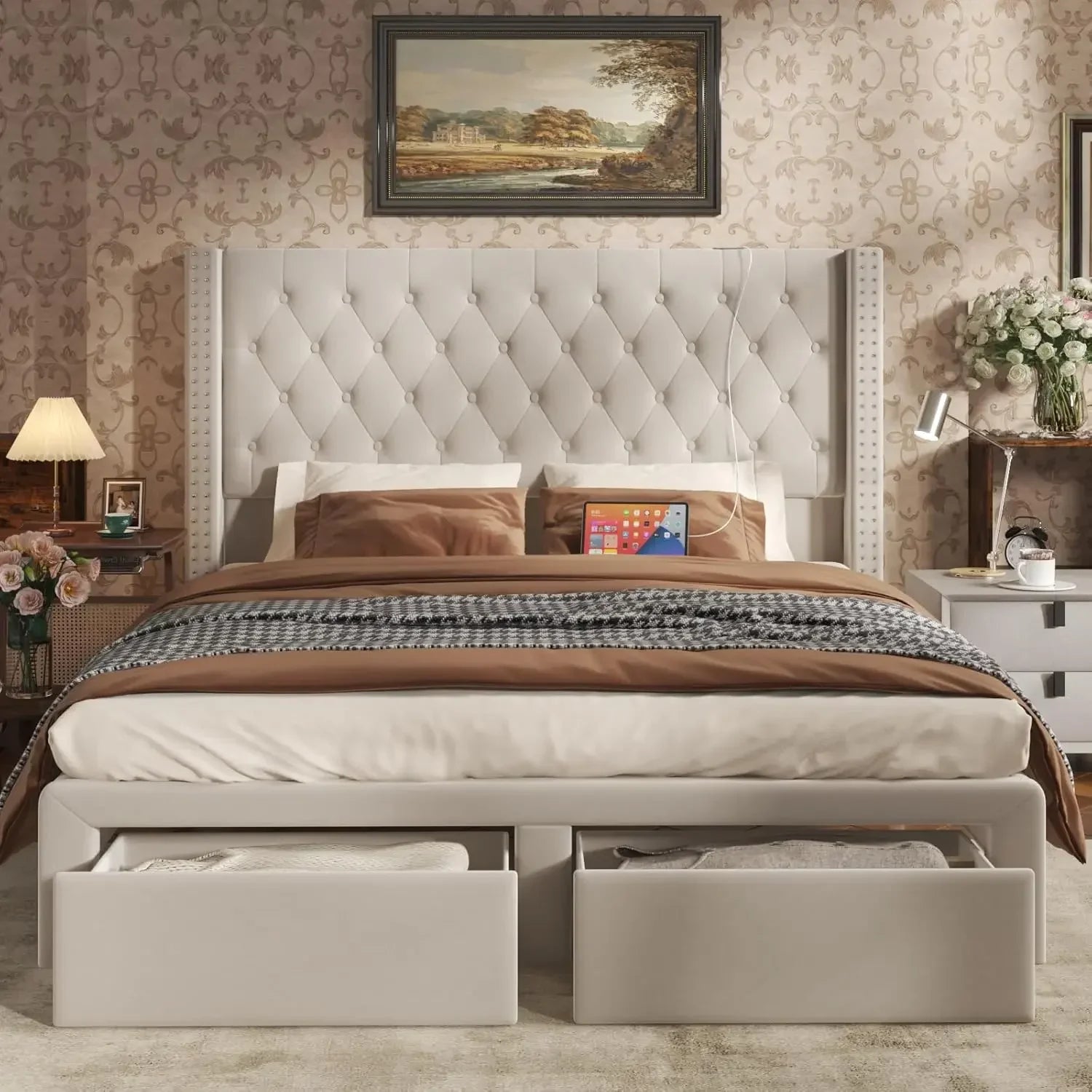 The Gio Velvet Queen Bed with Storage by Morsale creates a luxurious bedroom with its modern tufted headboard. Brown and beige cotton bedding complements elegant patterned wallpaper, while two storage drawers, a painting above, and bedside tables with lamps and flowers enhance the cozy ambience.