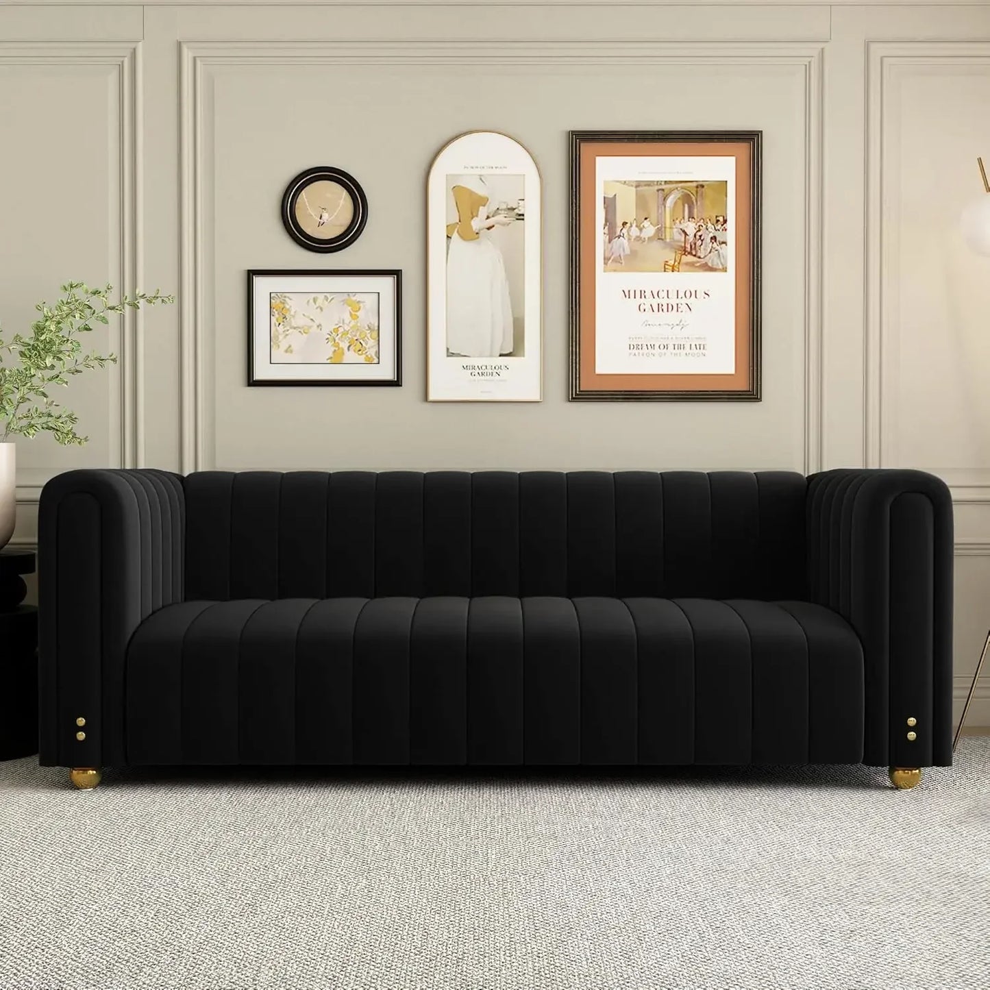 An 80" velvet living room sofa by Morsale, in a black Chesterfield style, sits against a light gray wall with artworks and a clock. A floor lamp stands to the right, while a small potted plant rests on the left. The setup exudes elegance and minimalism.