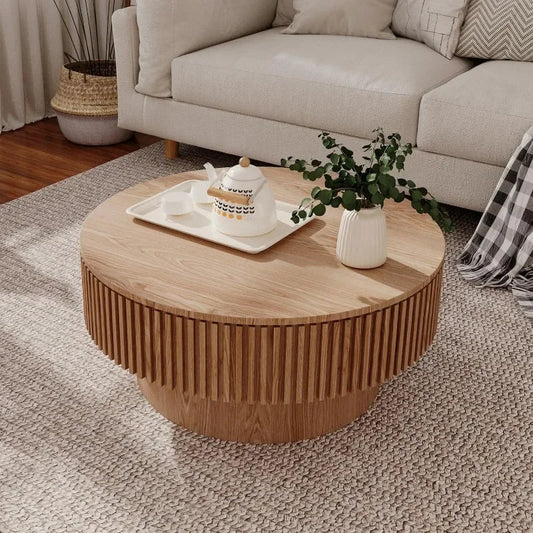 A Morsale 31" Round Coffee Table with Storage featuring a fluted design sits on a beige rug. It holds a teapot set on a tray and a vase of green foliage, while a cozy beige sofa and woven basket planter are in the background.