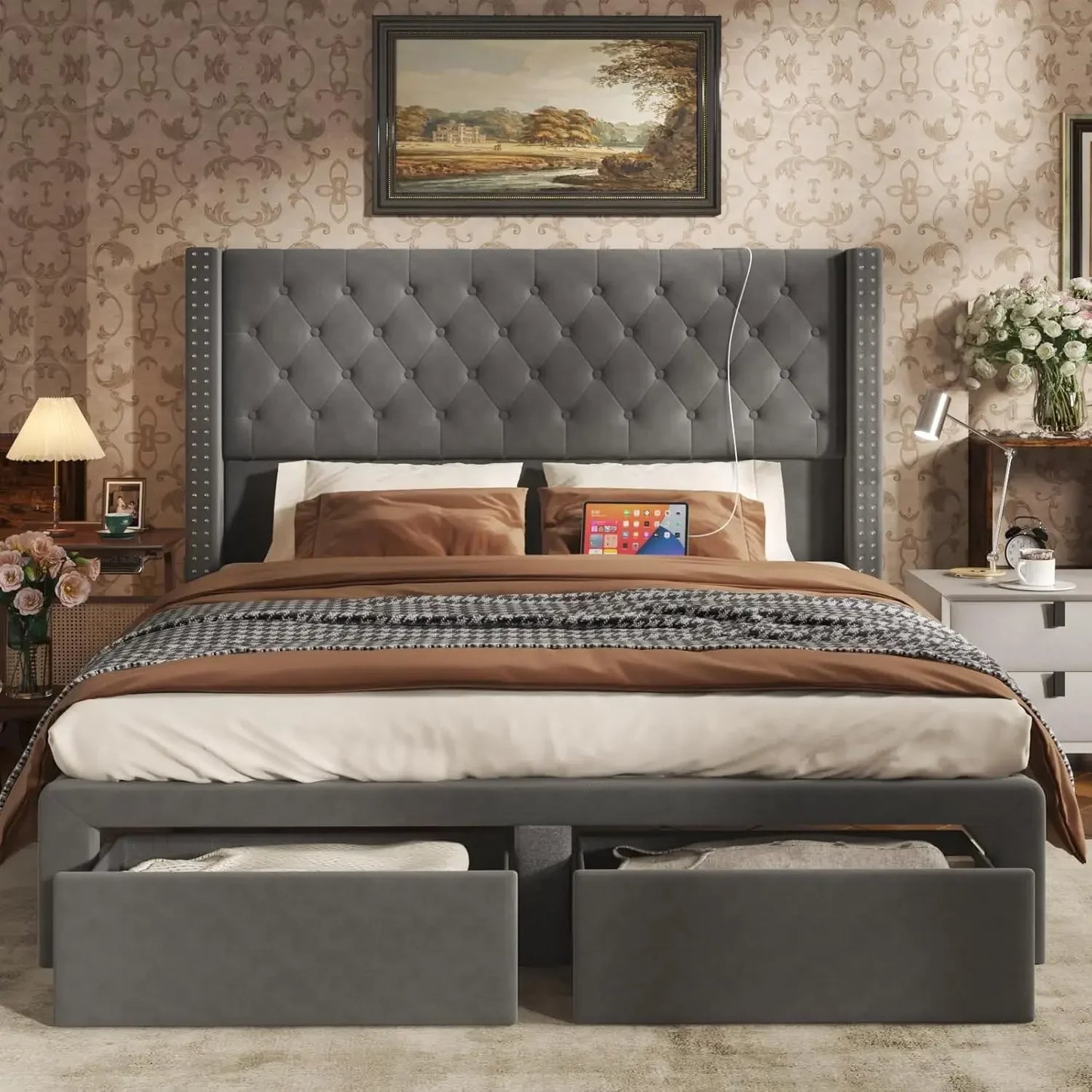 A bedroom showcases the Morsale Gio Velvet Queen Bed with a tufted gray headboard and built-in storage. It features brown and cream bedding, nightstands with lamps and flowers, subtle floral patterned wallpaper, and convenient built-in charging ports.