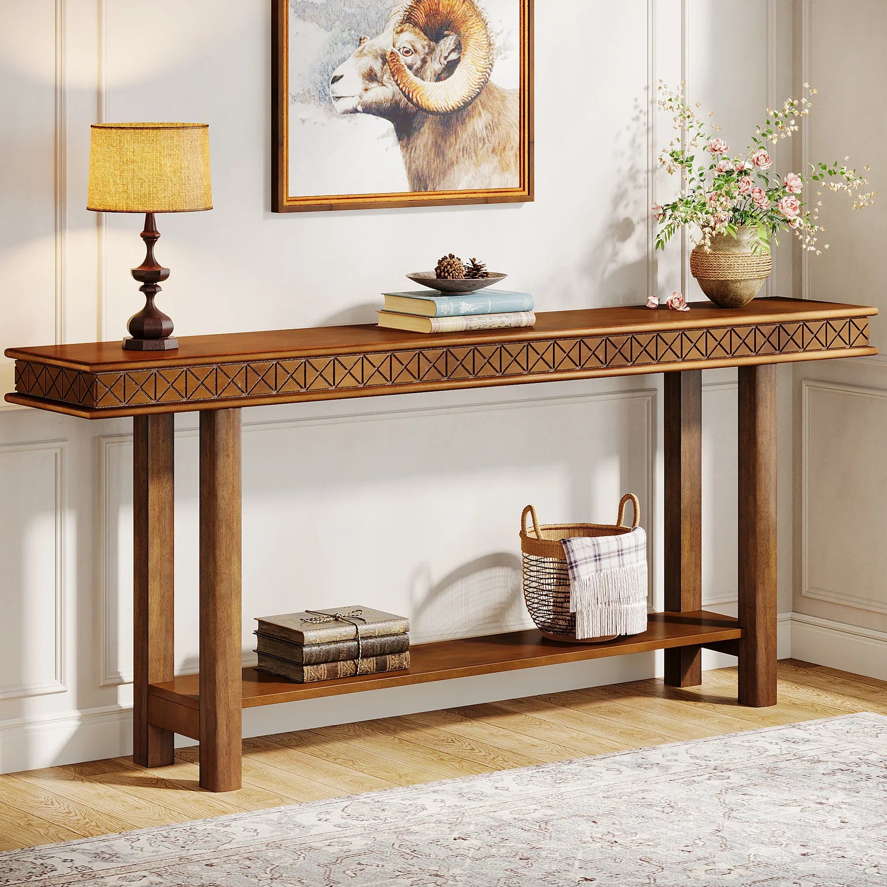 The Stanton Entryway Table With Storage by Morsale stands against a geometric-patterned wall, adorned with a lamp, books, and a vase of flowers. Below, a woven basket and more books sit on the lower shelf. A framed ram picture hangs above in this stylish piece of living room furniture with light walls and wooden flooring.