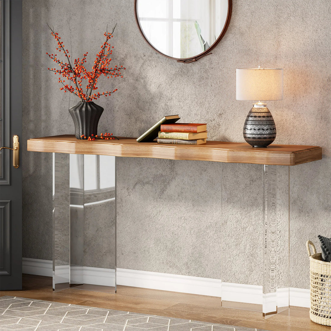 In a modern hallway, the Martin 47" Entryway Table with clear acrylic legs by Morsale displays books, a black vase with red berries, and a patterned lamp. A round mirror decorates the textured gray wall above. Nearby, a woven basket complements this chemical-free décor inspired by Mainland China craftsmanship.
