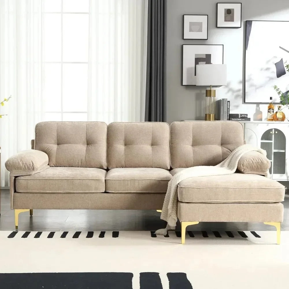 The Morsale 83" Sectional Sofa with Chaise in beige, featuring tufted cushions and gold legs, is complemented by a gray throw blanket. It harmonizes with white sheer curtains, a dark accent wall, framed art, and a white side cabinet decorated with lamps and ornaments.