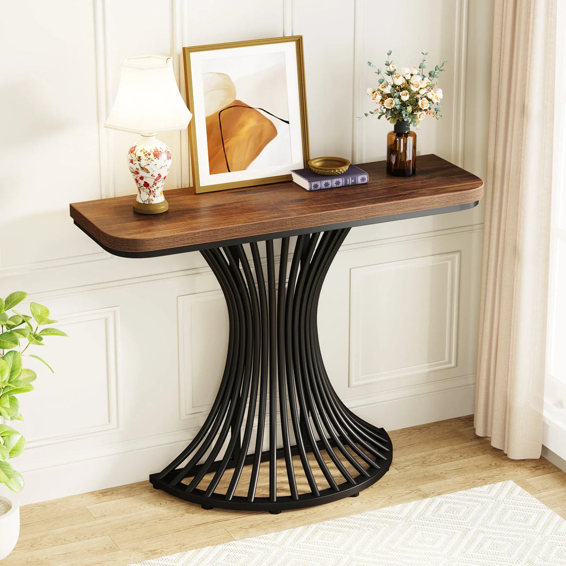 The Hinton 39-Inch Entryway Table by Morsale has a curved black metal base and wooden top with chemical-free finishes, displaying two ceramic jars, books, a small sculpture, and a plant. A round mirror hangs above it on the white paneled wall.