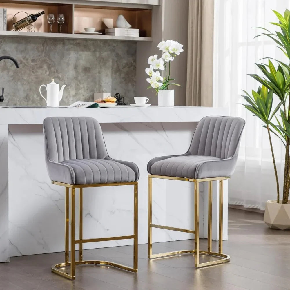 In a modern U.S.-style kitchen, a marble island features two gray velvet Modern Counter Height Bar Stools by Morsale with gold legs. A white teapot and potted orchid sit atop the counter. Nearby, a tall plant and open shelves showcase plates and cups in natural light.