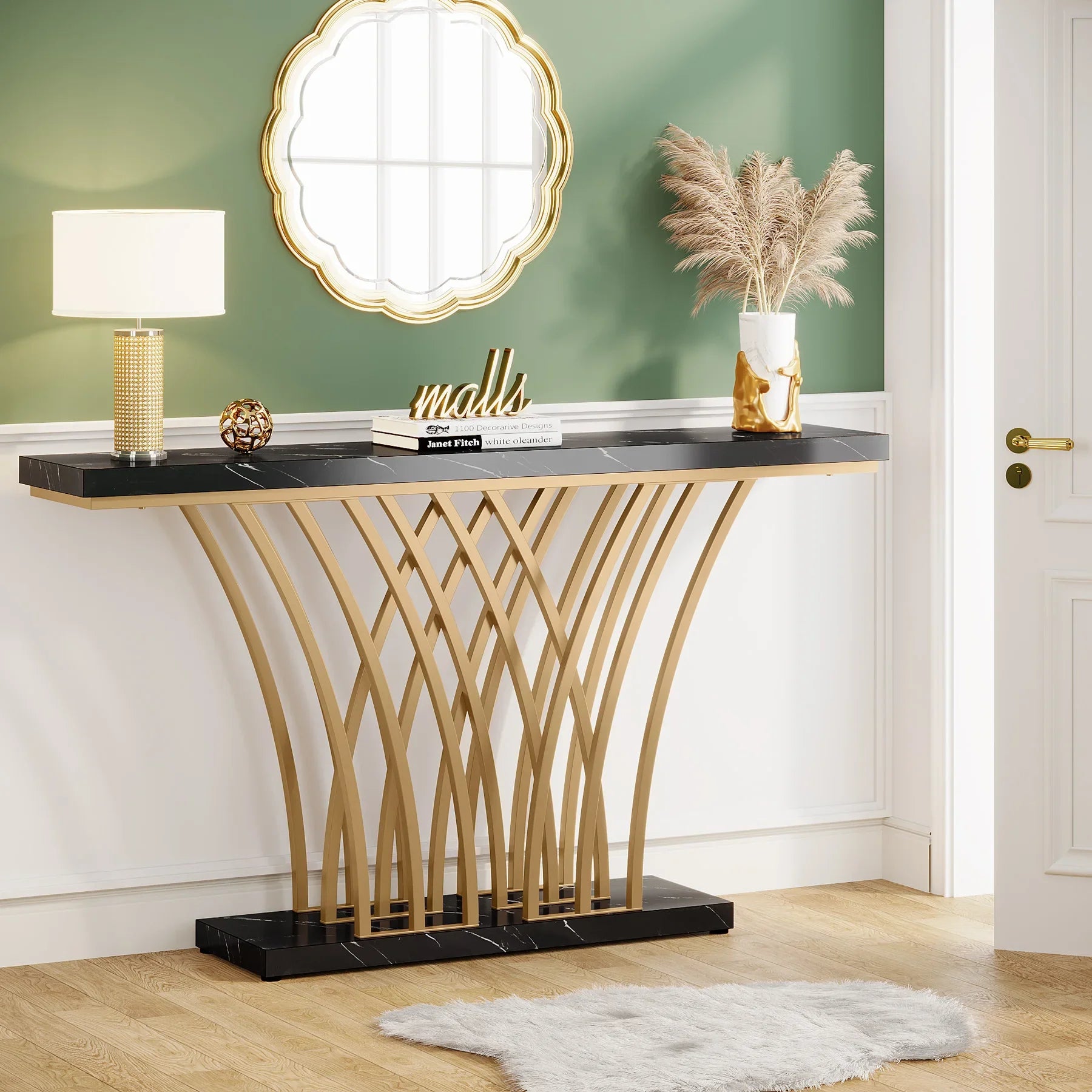 A modern entryway features the Bassett Entryway Table by Morsale, with a sleek black faux marble top and geometric gold metal base. It holds a lamp, decorative sphere, "malls" sign, and pampas grass in a vase beneath a round gold-framed mirror on the green wall. A light rug finishes the look.
