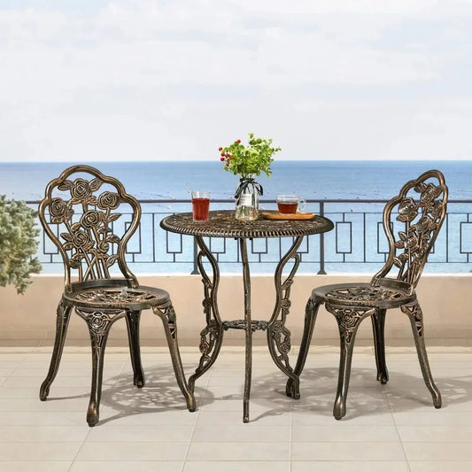 On the patio, the Davids 3-Piece Vintage Outdoor Set by Morsale features two ornate metal chairs and a round table with drinks and a plant. The area is bordered by a decorative railing, enhancing the tranquil seaside ambiance. The background showcases Morsale's stunning ocean view under a clear sky.