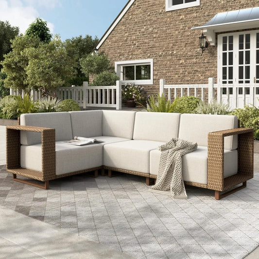 An outdoor patio features the modern Morsale Burton Outdoor Rattan Wicker Sofa Sectional with beige cushions and a knitted throw. It sits on a patterned tile patio surrounded by greenery, in front of a brick house with white trim and a picket fence.