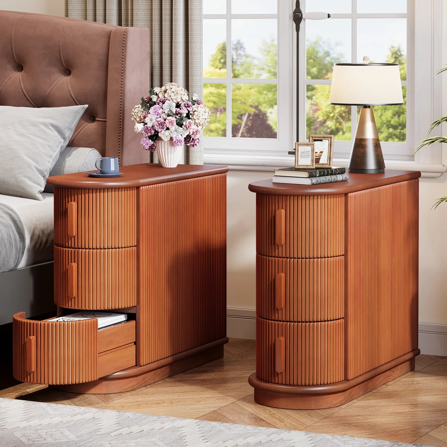 The Morsale Irvine Solid Wood Side Tables, featuring a ribbed design and vintage finish, flank the bed with a tufted headboard. One oak table's open drawer holds a blue cup, while the other supports a lamp, framed photos, and flowers. A window showcases greenery in the background.