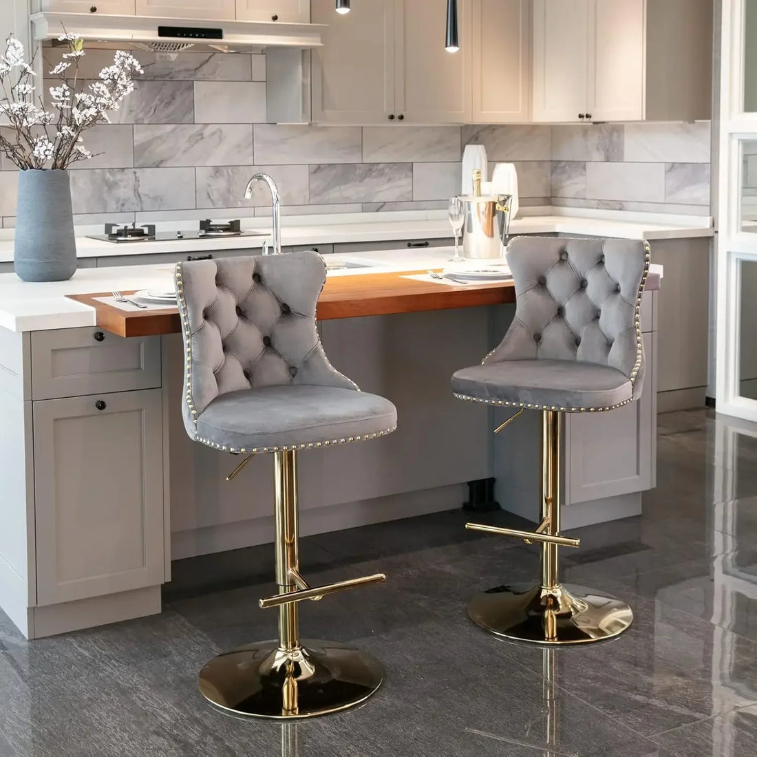 A modern kitchen features a white island with a wooden countertop, two ergonomic Morsale Counter Height Swivel Bar Stools in gray tufted designs and gold bases, complemented by white cabinets, stainless steel appliances, and an elegant vase with branches.