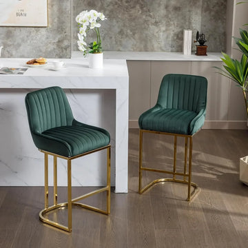 A set of two "Modern Counter Height Bar Stools" by Morsale, featuring green upholstery and a gold metal frame, complements a white marble kitchen island. A potted plant, an orchid on the counter, and a light gray textured wall complete the inviting atmosphere.