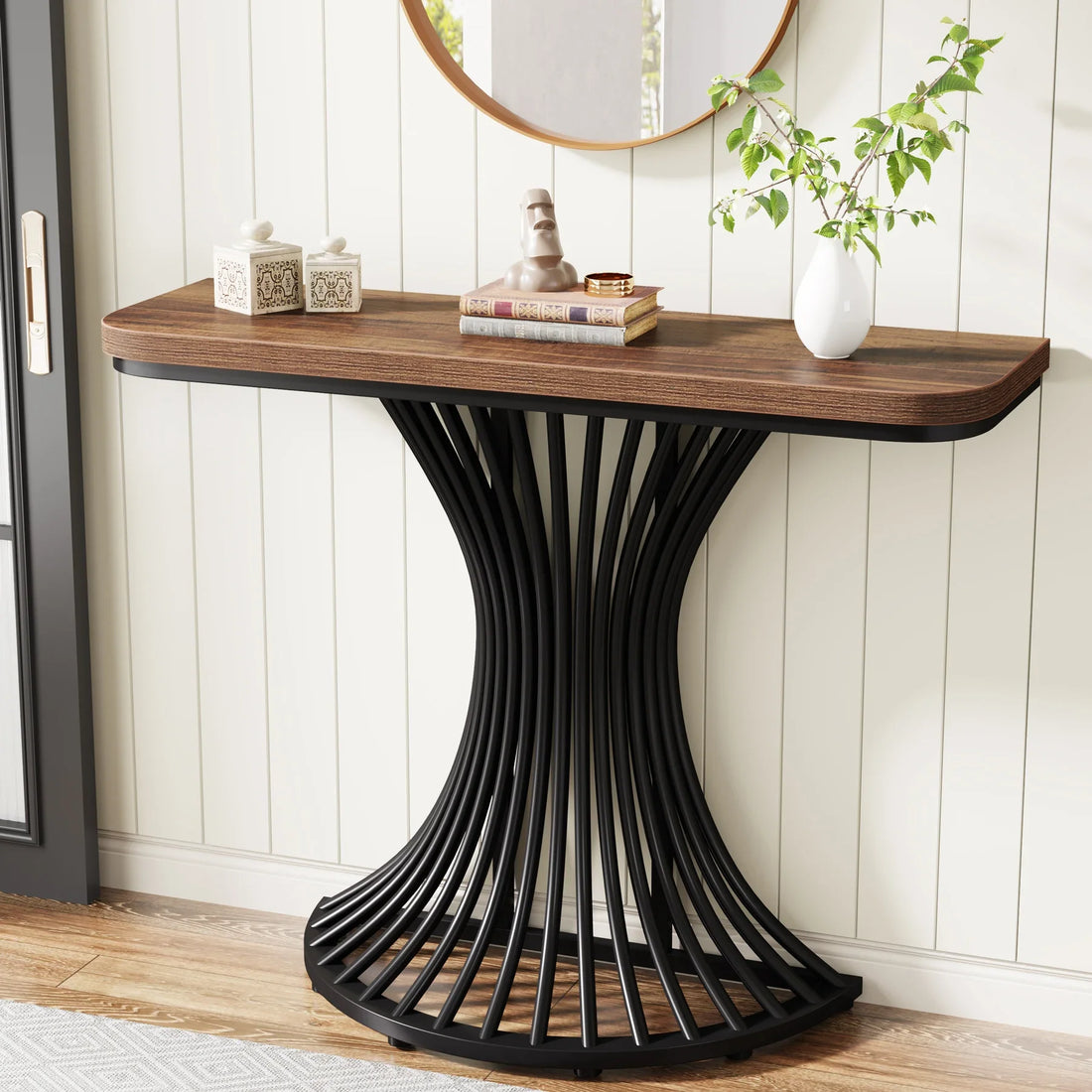The Hinton 39-Inch Entryway Table by Morsale has a curved black metal base and wooden top with chemical-free finishes, displaying two ceramic jars, books, a small sculpture, and a plant. A round mirror hangs above it on the white paneled wall.