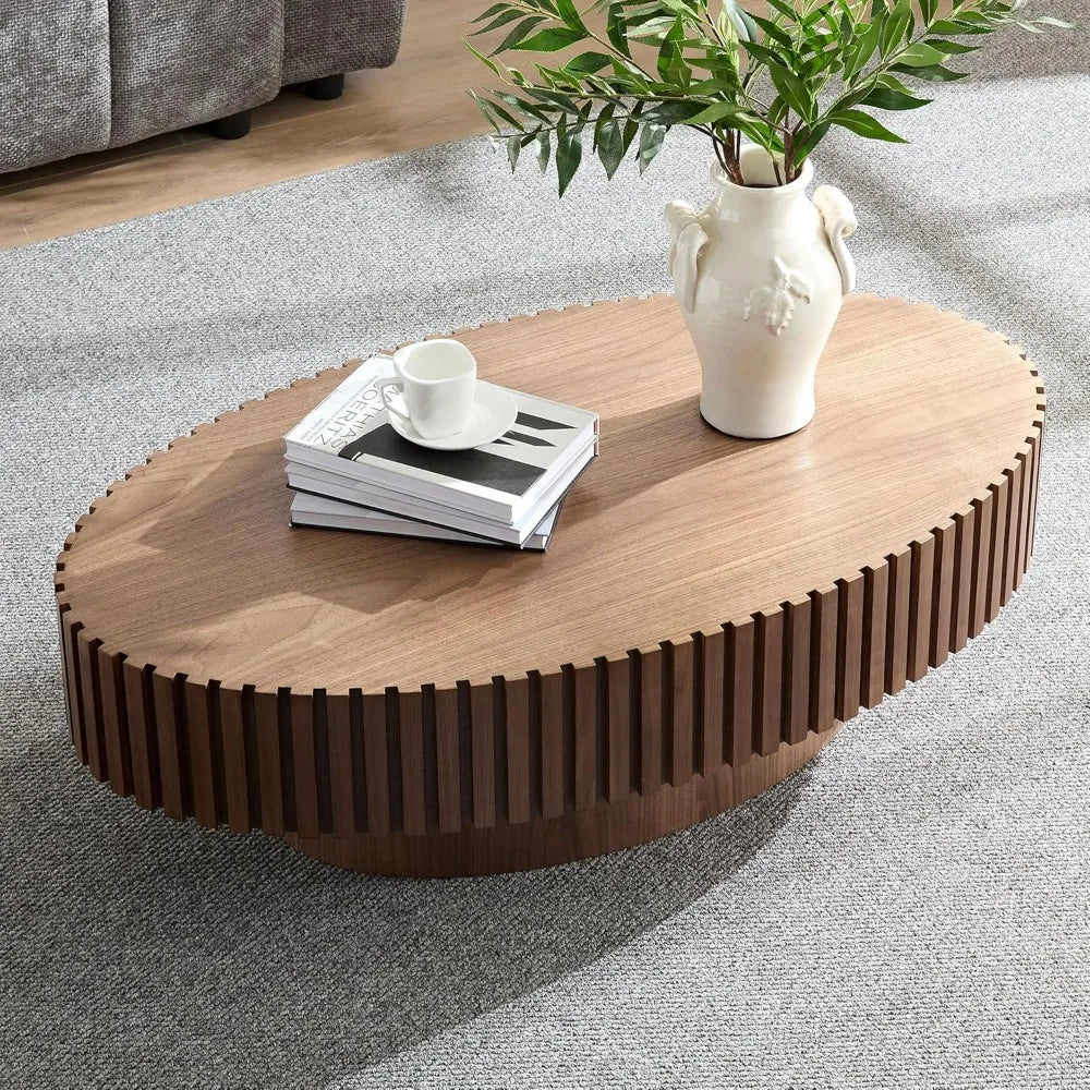 The Morsale 43" oval wooden coffee table features a textured edge with a white vase, plant, books, and cup on top. Placed on a gray carpeted floor with a couch in the background, this chemical-free piece offers style and safety for your living room.
