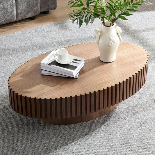 The Morsale 43" oval wooden coffee table features a textured edge with a white vase, plant, books, and cup on top. Placed on a gray carpeted floor with a couch in the background, this chemical-free piece offers style and safety for your living room.