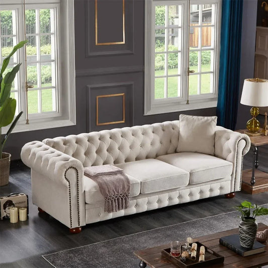 The living room showcases an elegant 3-Seat Chesterfield Sofa by Morsale, accented with a throw blanket and pillow. Dark wood flooring, potted plant, and large windows with a garden view complement the decor, which includes candles and a glass table for an inviting, stylish feel.