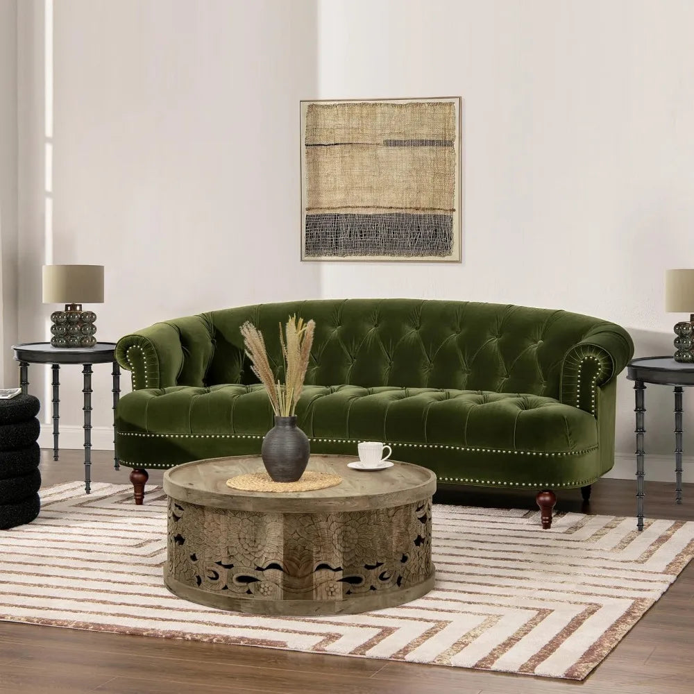 A living room includes a Chesterfield Velvet Tufted Sofa in Olive Green by Morsale, a round wooden coffee table, two side tables with matching lamps, and is adorned with a neutral striped rug. A textured abstract art piece enhances the elegance of the light-colored walls.