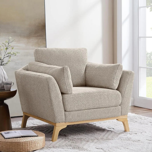 The Morsale Sofi Living Room Accent Chair, a cozy beige masterpiece with wooden legs, complements the patterned rug and bright living room. A cylindrical cushion graces the chair. Nearby, a side table with a potted plant and cup sits in view of the large window.