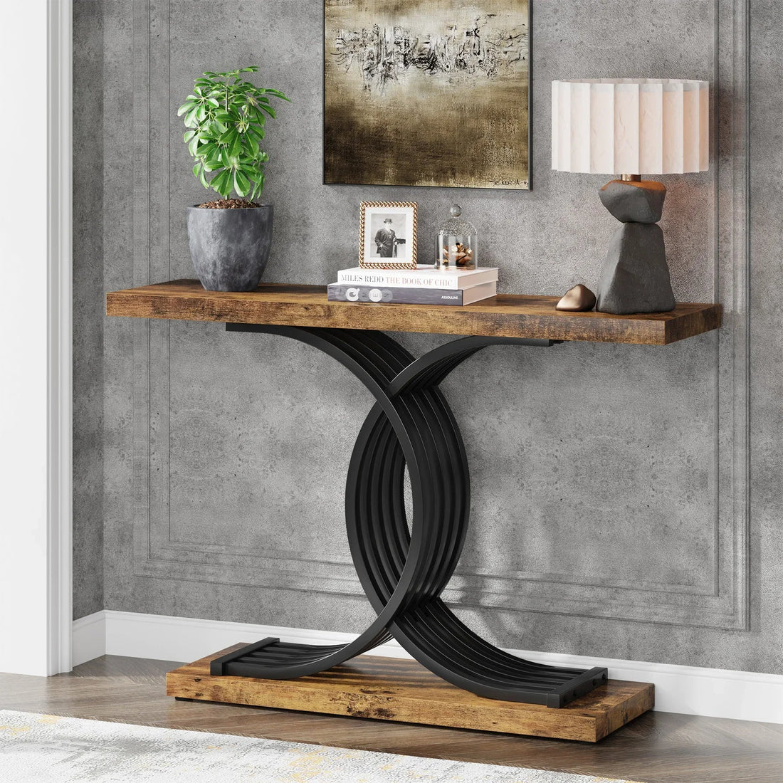 The Morsale Wexford Geometric Frame Console Table, featuring a decorative black metal base, stands against a textured gray wall with a lamp, potted plant, and books on top. Above this modern-style wooden table hangs a framed abstract painting.