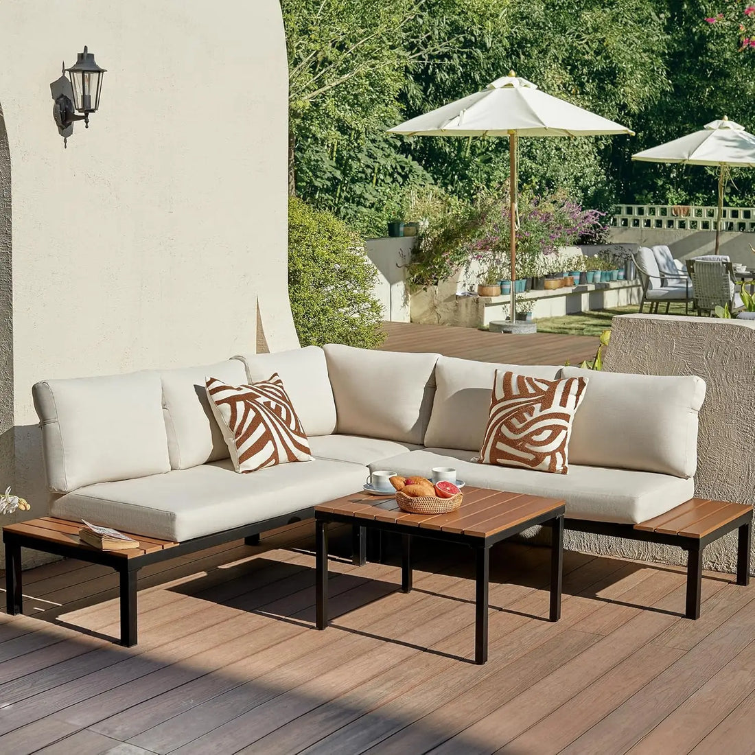 An outdoor patio featuring the Warton Patio Set by Morsale, with a beige all-weather sectional sofa, two patterned pillows, and a wooden coffee table holding snacks and drinks. Umbrellas and greenery create a serene garden ambiance.